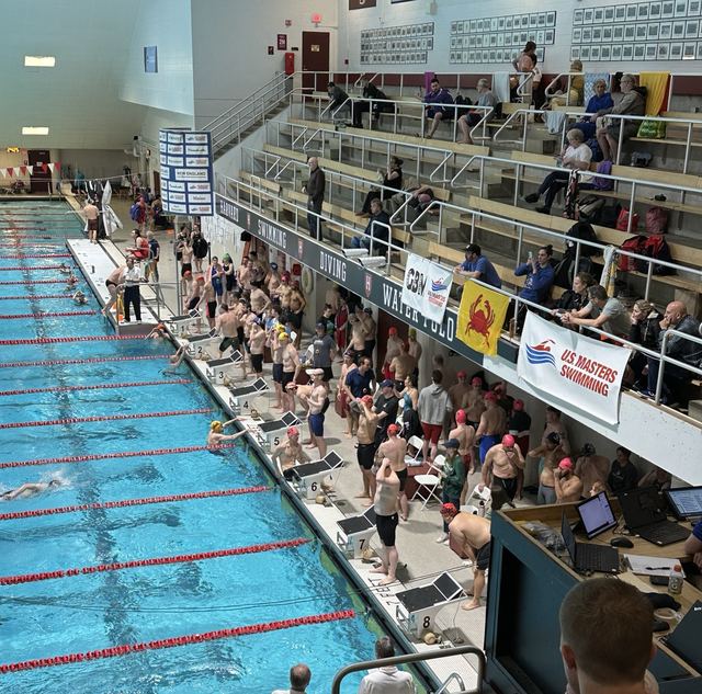 Masters swimmers waiting behind the starting blocks along a competition pool, with a US Masters Swimming banner hanging above the deck.